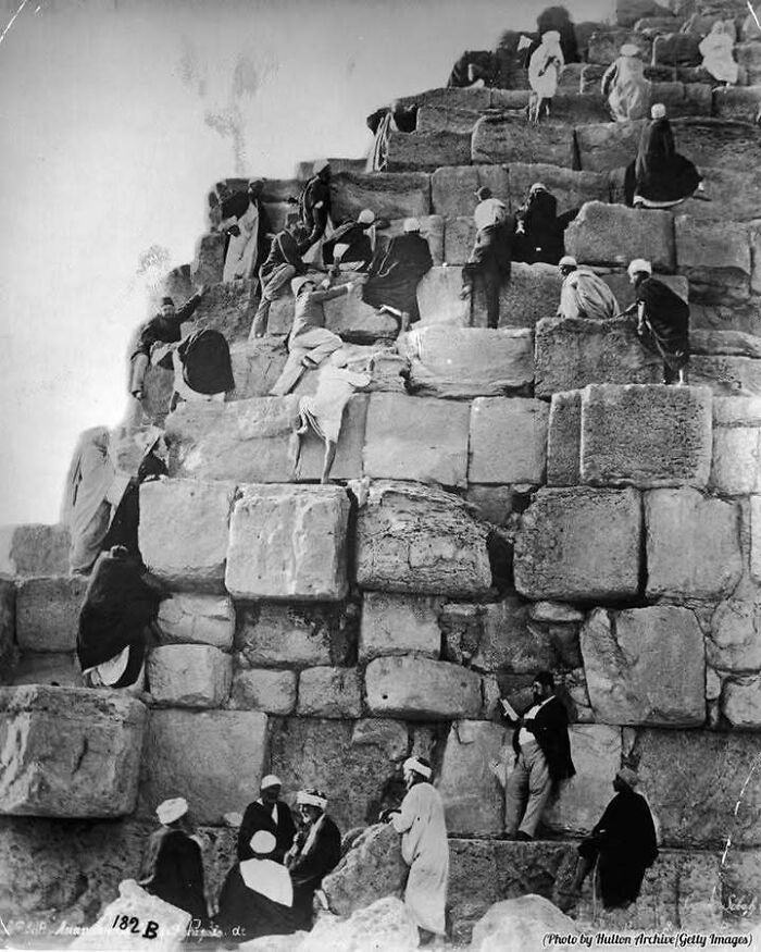 People climbing and sitting on ancient stone blocks of a classic historical structure in a black and white photo.
