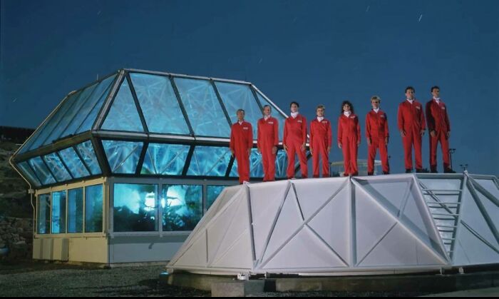Group of people in red suits standing on a modern structure under a night sky in a classic historical picture setting