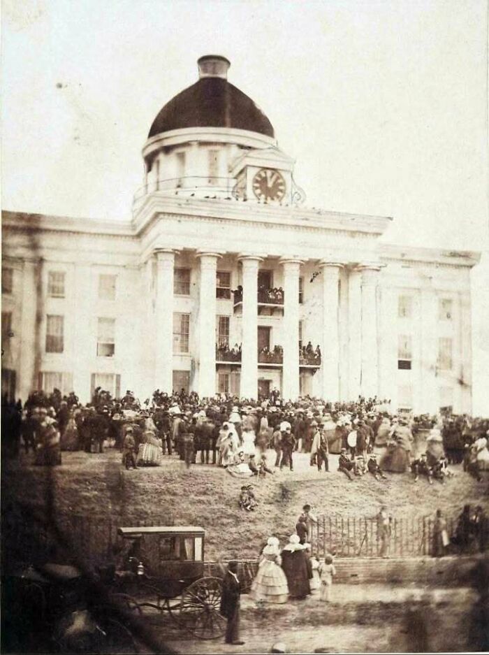 Historic sepia photo of a large crowd gathered at a classic building with columns and a clock tower, iconic historical picture.