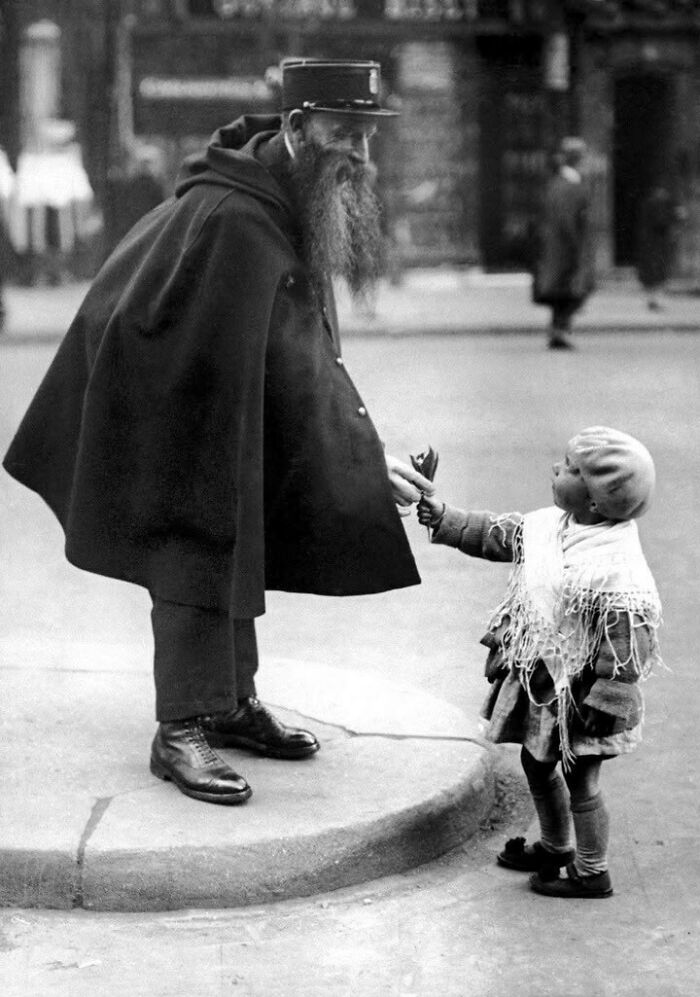 Black and white historical picture of an elderly man in uniform receiving a flower from a young child on the street.