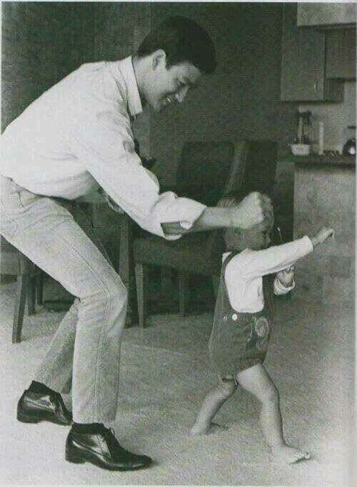 Black and white historical picture of a man helping a toddler take first steps indoors, classic family moment captured.
