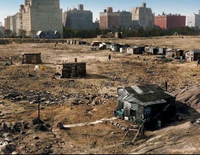 Shantytown with makeshift homes and barren land in front of classic city buildings in a historical picture.