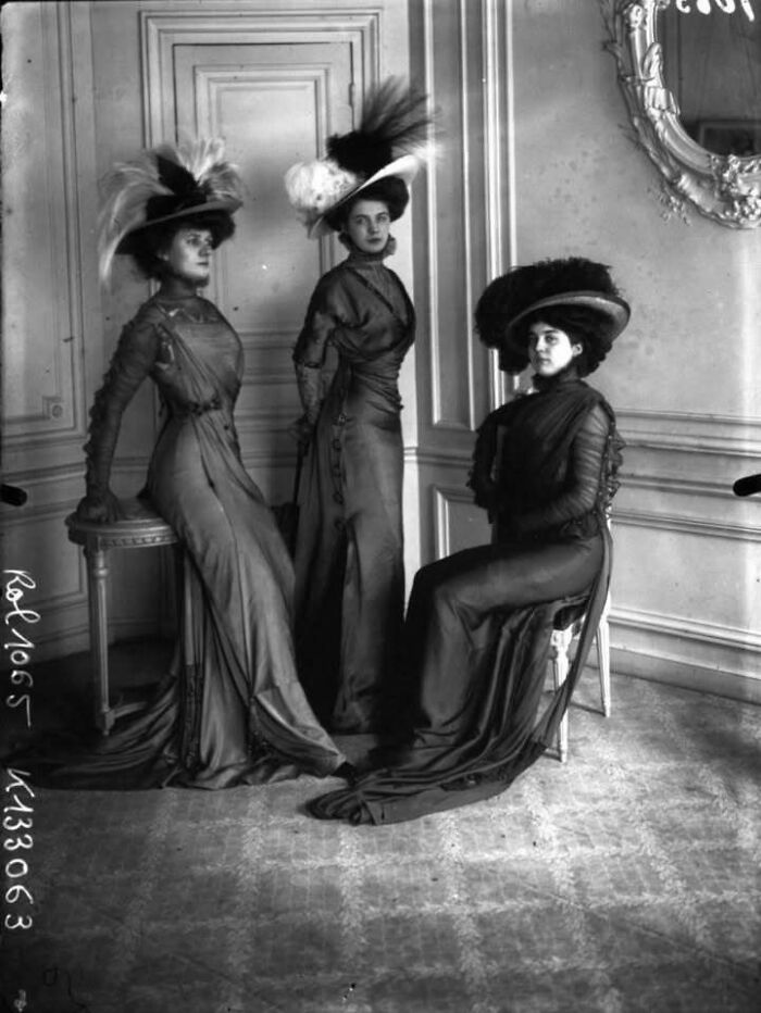 Three women in elegant vintage dresses and large feathered hats posing inside a classic historical setting.