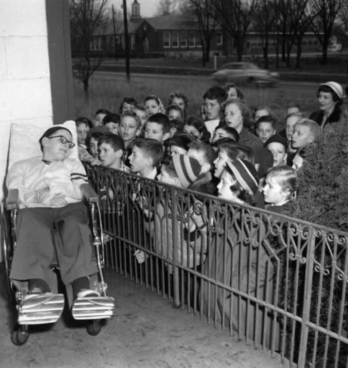 Black and white historical picture of children gathered around a man in a wheelchair sharing a moment outdoors.