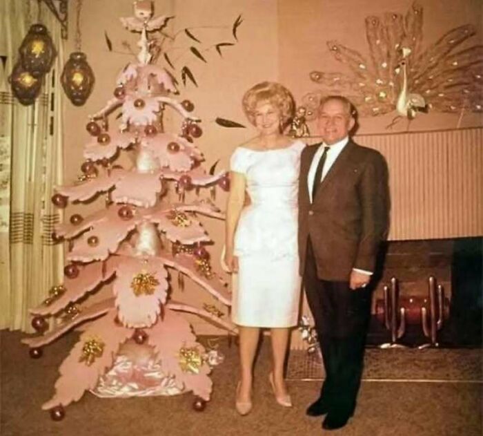 Vintage couple posing beside a uniquely decorated pink Christmas tree in a classic historical picture from the 1960s.