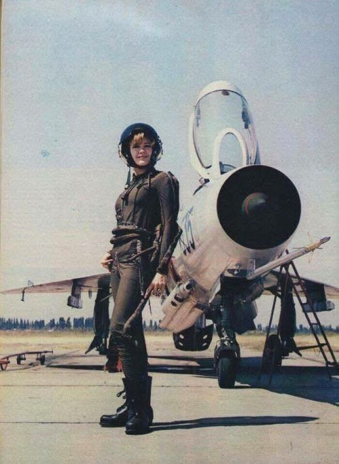 Young female pilot in flight suit stands confidently next to a military jet in a classic historical picture.