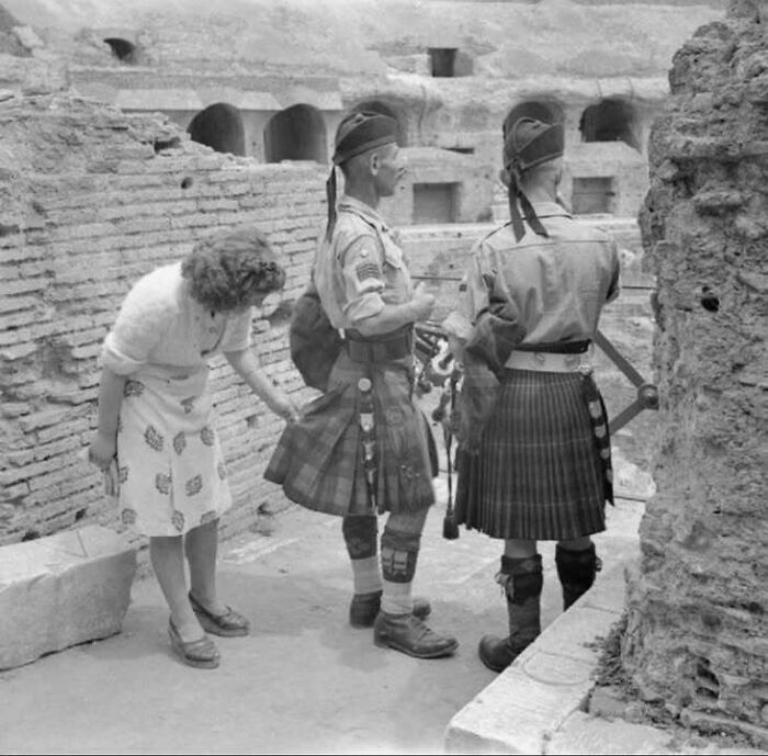 Two soldiers wearing kilts and a woman examining one kilt amid ancient ruins in a classic historical picture.