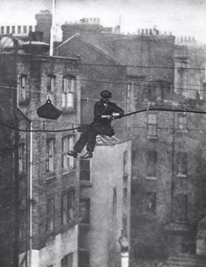 Man performing high-wire act above city streets in a classic black and white historical picture.