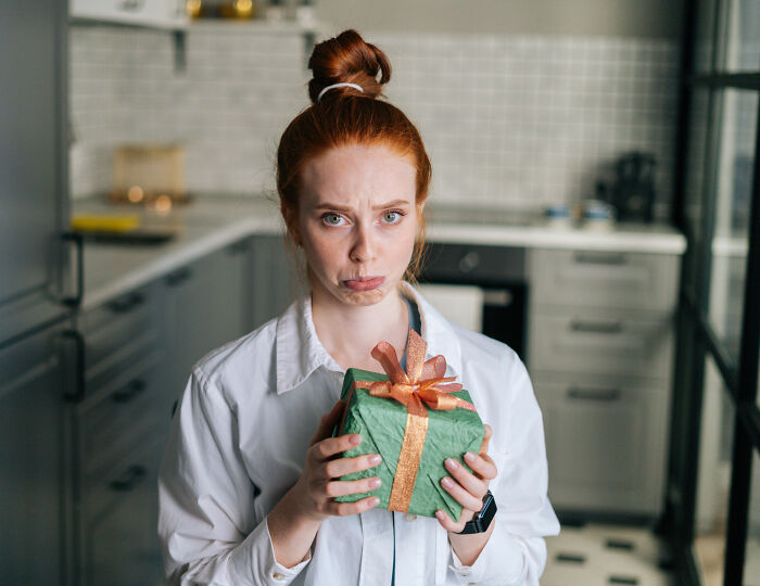 Red-haired woman holding holiday gift with a sad expression, suggesting painful truth about relationship and partner’s gift. Red-haired woman holding holiday gift with a sad expression, suggesting painful truth about relationship and partner’s gift.