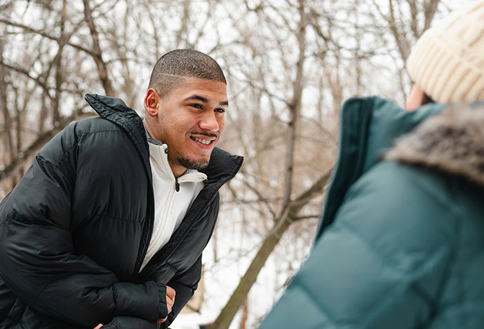 A young man in a black jacket smiling outdoors in winter, with trees and snow in the background.