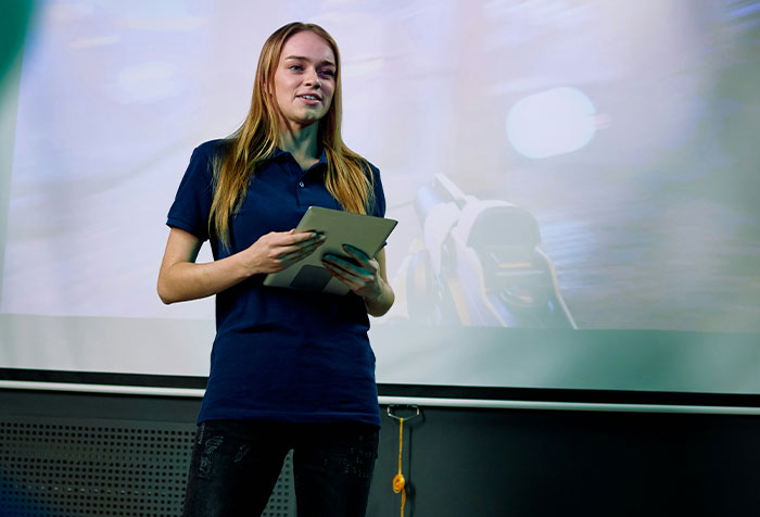 A young woman holding a tablet, speaking confidently in front of a screen, embodying acts of microfeminism.