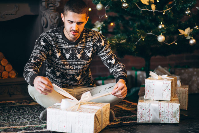 Young man in festive sweater sitting by Christmas tree, unwrapping presents, reflecting on pregnancy and baby plans before 30.