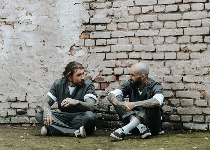 Two inmates with tattoos sitting on the ground against a brick wall discussing prison life facts and misconceptions.
