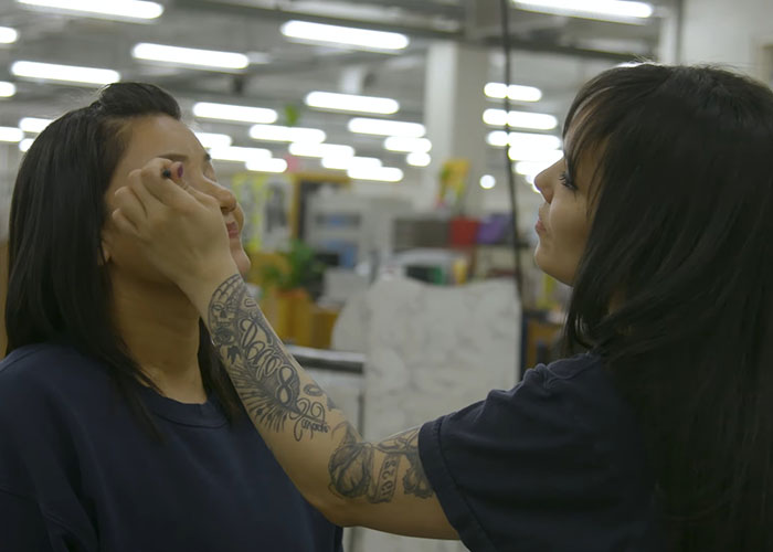 Two inmates in prison uniforms inside a facility, one applying makeup to the other, showing prison life realities.