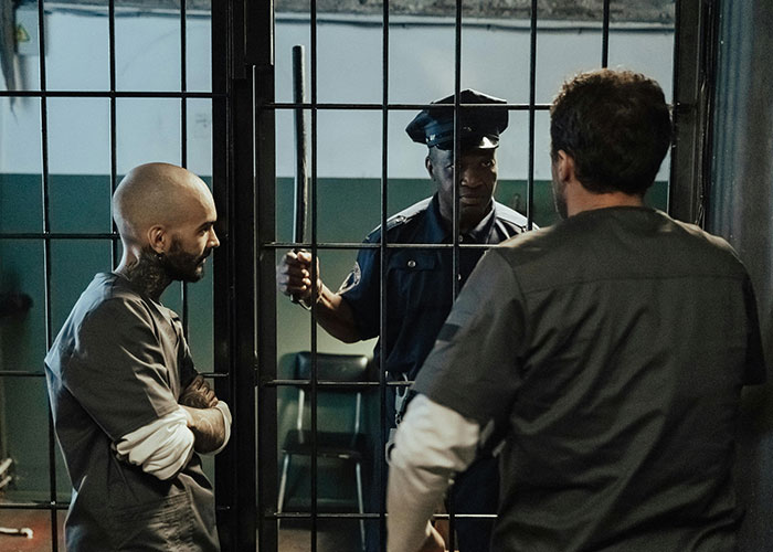 Two inmates in prison uniforms talking to a guard through bars illustrating prison life misconceptions.