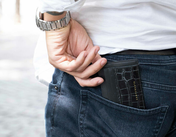 Man wearing a silver watch putting a black wallet in the back pocket of dark blue jeans outdoors.