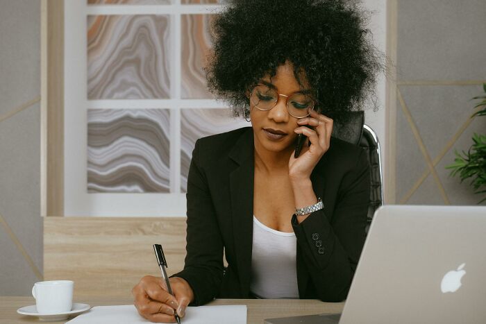 Woman with curly hair and glasses making notes while talking on the phone, illustrating quitting jobs in record time stories.
