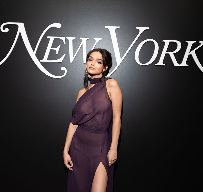 Rachel Zegler wearing a purple see-through dress with a high slit posing in front of New York sign on a black background. Rachel Zegler wearing a purple see-through dress with a high slit posing in front of New York sign on a black background.