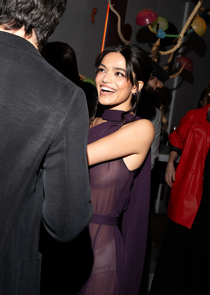 Rachel Zegler smiling in a purple see-through dress at an event, engaging happily with a guest in a dark jacket. Rachel Zegler smiling in a purple see-through dress at an event, engaging happily with a guest in a dark jacket.