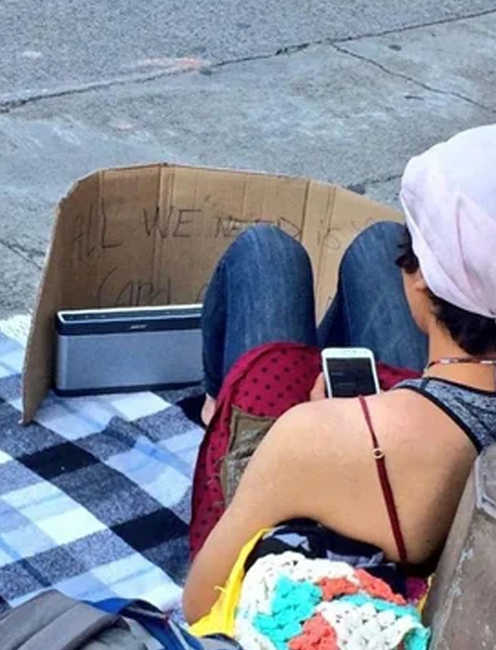 Person sitting on a sidewalk with a cardboard sign and speaker, a calm moment amid rage-inducing situations.