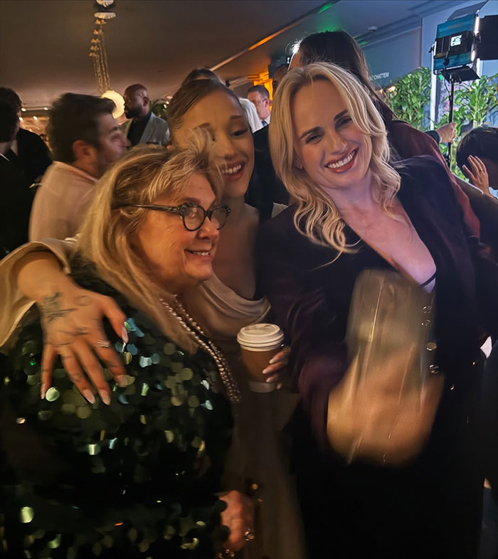Rebel Wilson and Ariana Grande smiling and posing for a selfie at a crowded indoor event with people in the background. Rebel Wilson and Ariana Grande smiling and posing for a selfie at a crowded indoor event with people in the background.