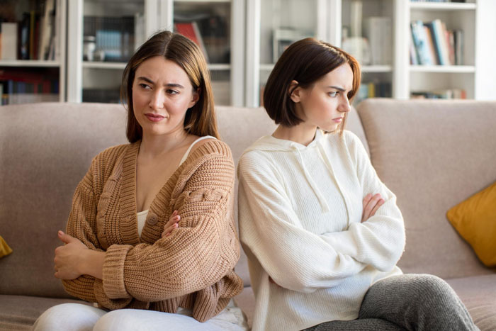Two women sitting back-to-back on a couch with arms crossed, showing tension over family heirloom ring conflict.