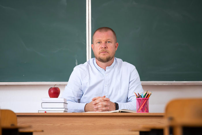 Man in light blue shirt sitting at desk in classroom, appearing tense and suspicious of younger colleague's intentions. Man in light blue shirt sitting at desk in classroom, appearing tense and suspicious of younger colleague's intentions.