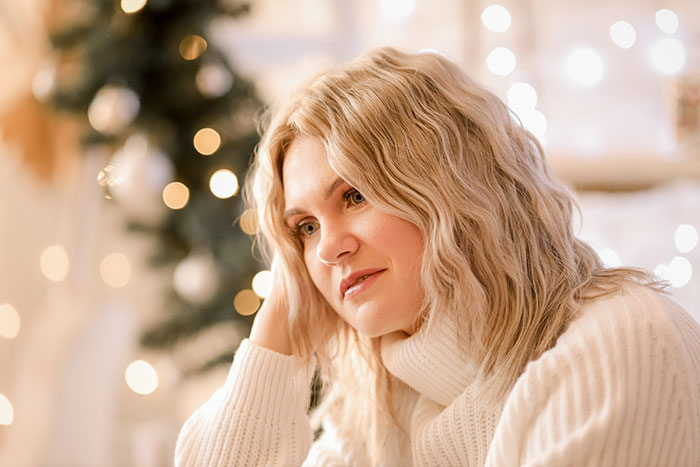 Woman in white sweater looking thoughtful near Christmas tree with lights, reflecting on hosting Christmas dinner demands. Woman in white sweater looking thoughtful near Christmas tree with lights, reflecting on hosting Christmas dinner demands.