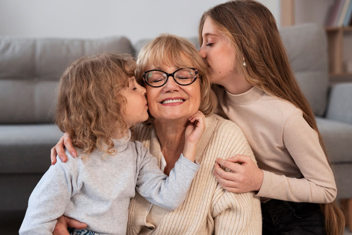 Grandmother receiving kisses from two children in a living room, symbolizing family bonds and affection. Grandmother receiving kisses from two children in a living room, symbolizing family bonds and affection.