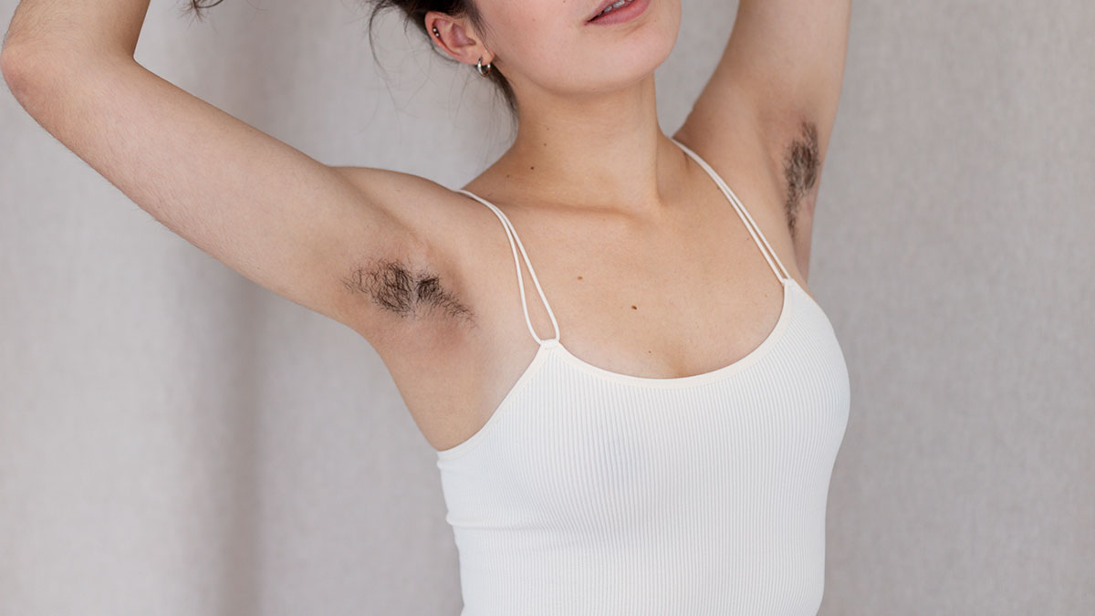 Woman showing hairy armpits while wearing a white tank top, highlighting natural body hair and personal grooming choice.