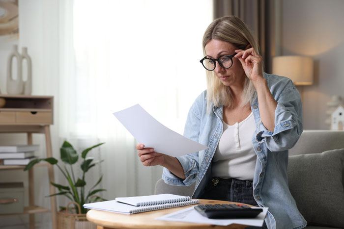 Woman reviewing HOA rules with glasses on, sitting at a table with papers and a calculator indoors. Woman reviewing HOA rules with glasses on, sitting at a table with papers and a calculator indoors.
