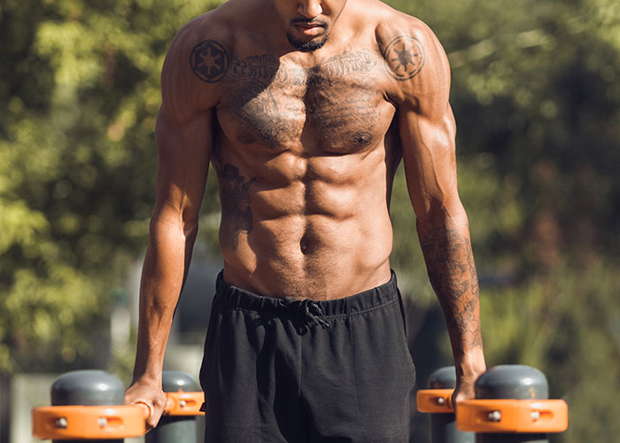 Muscular man outdoors doing parallel bar dips with focus on his bare chest and arms in natural light.