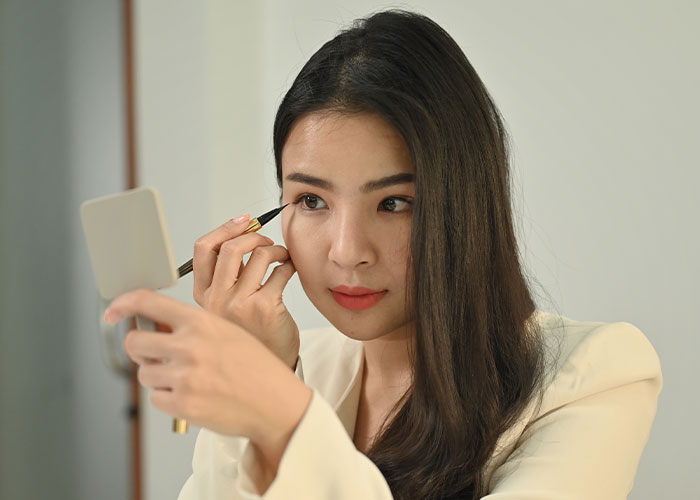 Young woman applying eyeliner while holding a mirror, illustrating makeup and the no makeup look discussion online. Young woman applying eyeliner while holding a mirror, illustrating makeup and the no makeup look discussion online.
