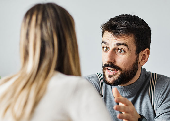 Man with beard talking animatedly to woman with long hair in a casual setting, illustrating online roasting of no makeup preference.