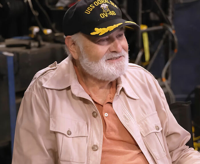 Bearded man wearing USS OORAL SEA baseball cap and beige jacket, smiling in an indoor setting discussing MAGA hypocrisy. Bearded man wearing USS OORAL SEA baseball cap and beige jacket, smiling in an indoor setting discussing MAGA hypocrisy.