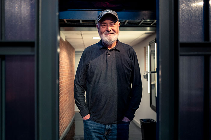 Rob Reiner standing casually in a hallway with hands in pockets, wearing a cap and dark long-sleeve shirt.