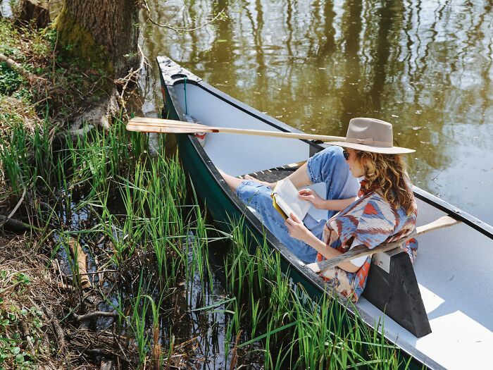 Woman wearing a hat and reading a book while sitting in a canoe near water surrounded by reeds, highlighting everyday health dangers.
