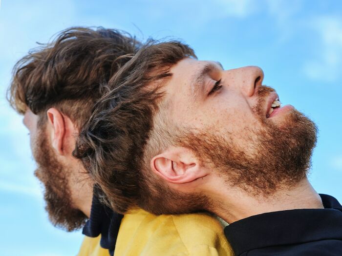 Two bearded men leaning back to back outdoors under a blue sky symbolizing incredible minds and genius.