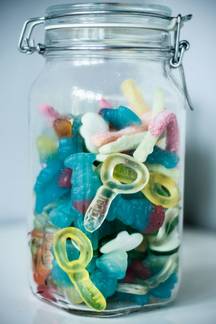 Glass jar filled with colorful gummy candies shaped like pacifiers, worms, and other playful forms.