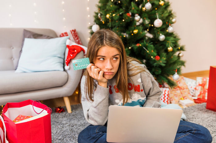 Young woman holding credit card, looking upset during family Secret Santa near Christmas tree and gifts at home. Young woman holding credit card, looking upset during family Secret Santa near Christmas tree and gifts at home.