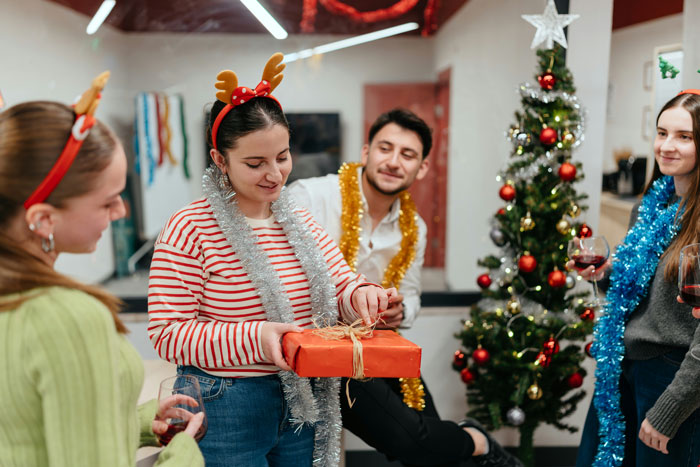Four friends celebrating Christmas with gift exchange and festive decorations including a decorated tree and tinsel accessories
