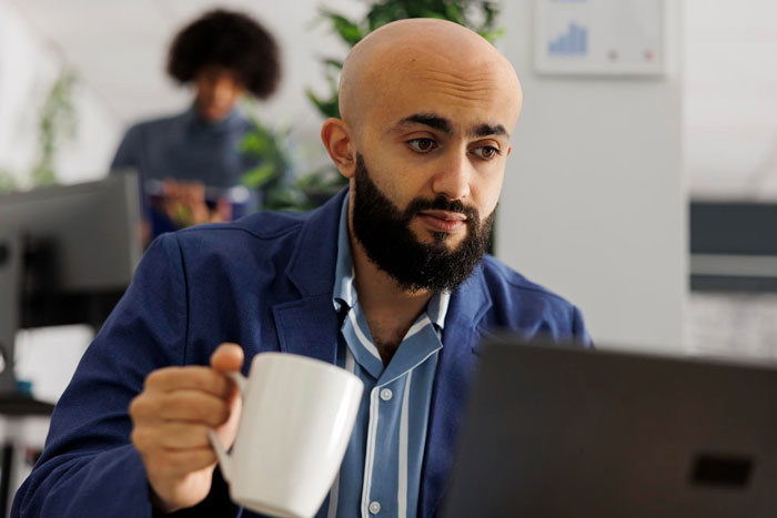 Man looking conflicted at laptop while holding a mug in an office, relating to coworker birth babies comment dilemma.