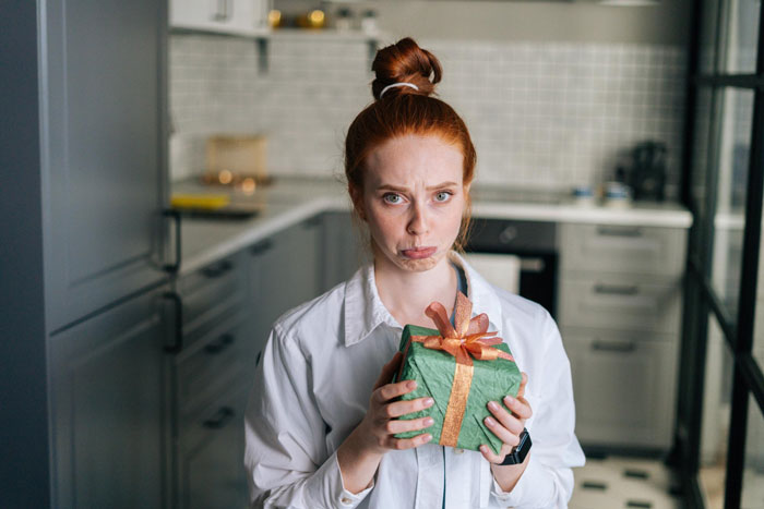 Jovem de cabelo ruivo segurando um presente de Natal, parecendo desapontada enquanto estava em uma cozinha moderna para uma lista de presentes de Natal. Jovem de cabelo ruivo segurando um presente de Natal, parecendo desapontada enquanto estava em uma cozinha moderna para uma lista de presentes de Natal.