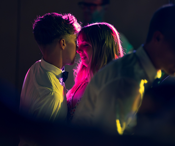 Young couple close together with the girl blushing while family members surround them at a dimly lit gathering. Young couple close together with the girl blushing while family members surround them at a dimly lit gathering.
