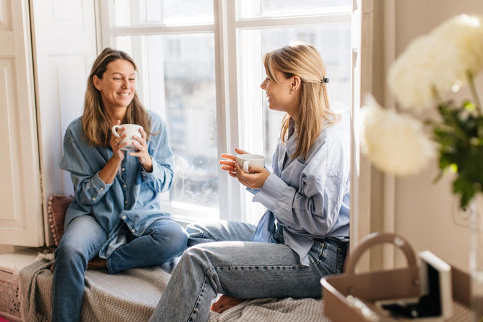 Two women sitting by a window, smiling and holding mugs, illustrating a story about a man lying to please fiancée. Two women sitting by a window, smiling and holding mugs, illustrating a story about a man lying to please fiancée.