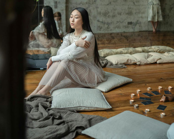 Young barefoot spiritual advisor sitting on floor cushions surrounded by candles and tarot cards in a calm room setting.