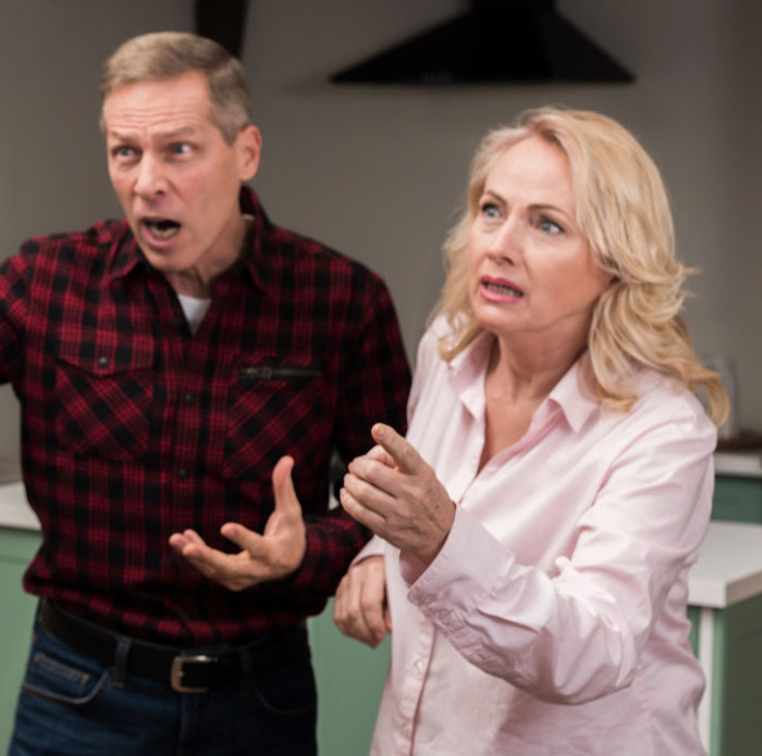 Couple arguing indoors, reacting with shock and frustration after sister’s barefoot spiritual advisor causes comical chaos.