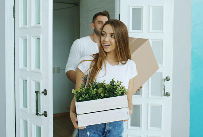 Young woman helping her brother and his wife by carrying a box of plants into their home showing family support and care.