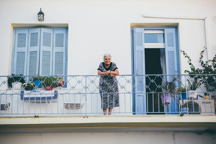 Elderly woman standing on balcony of traditional white Greek village house with blue shutters and plants. Elderly woman standing on balcony of traditional white Greek village house with blue shutters and plants.