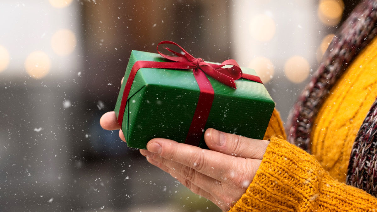 Person wearing a yellow sweater holding a green gift box tied with a red ribbon in falling snow outdoors, small tricks concept.